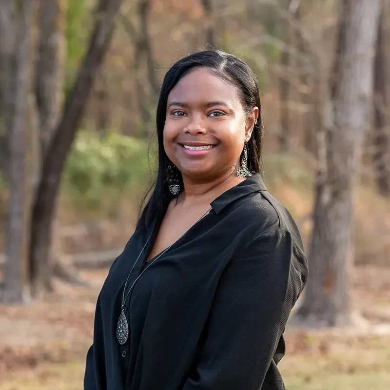 A smiling woman wearing a black top and a long necklace poses outdoors against a blurred, wooded background.