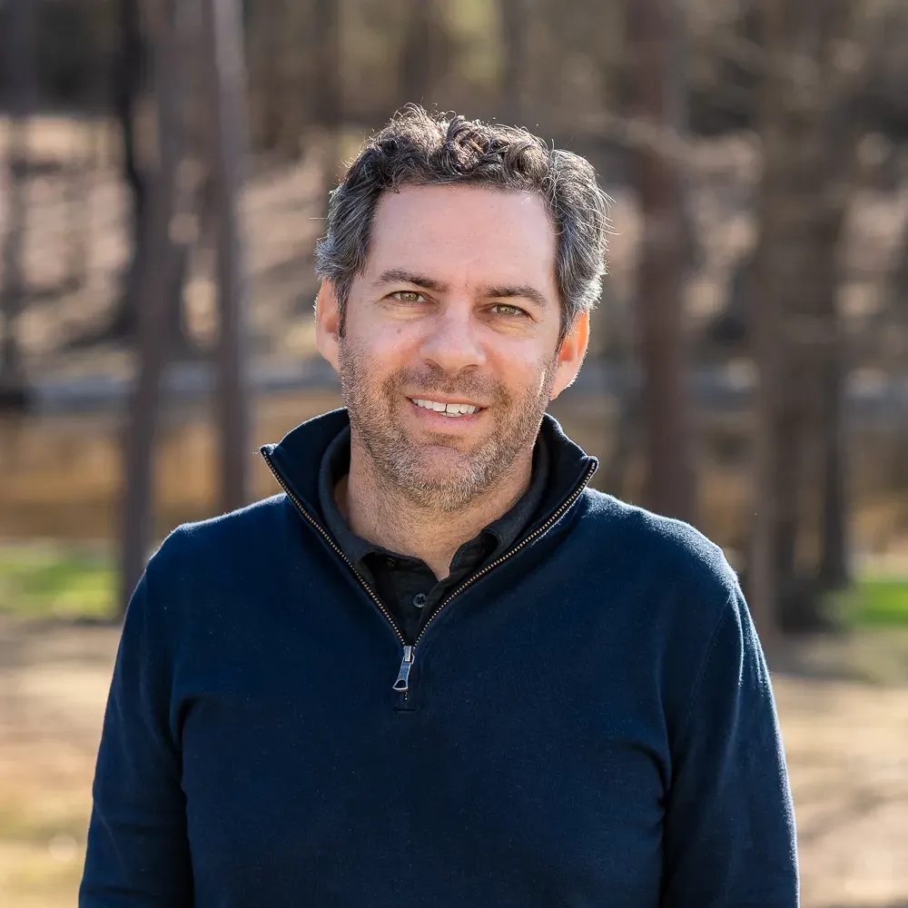 A person with short, dark curly hair and a beard smiles warmly, wearing a navy blue quarter-zip sweater in a park.