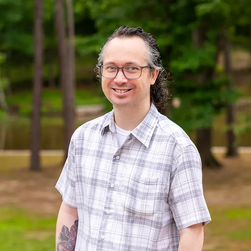 Smiling person in glasses and plaid shirt standing outdoors among trees