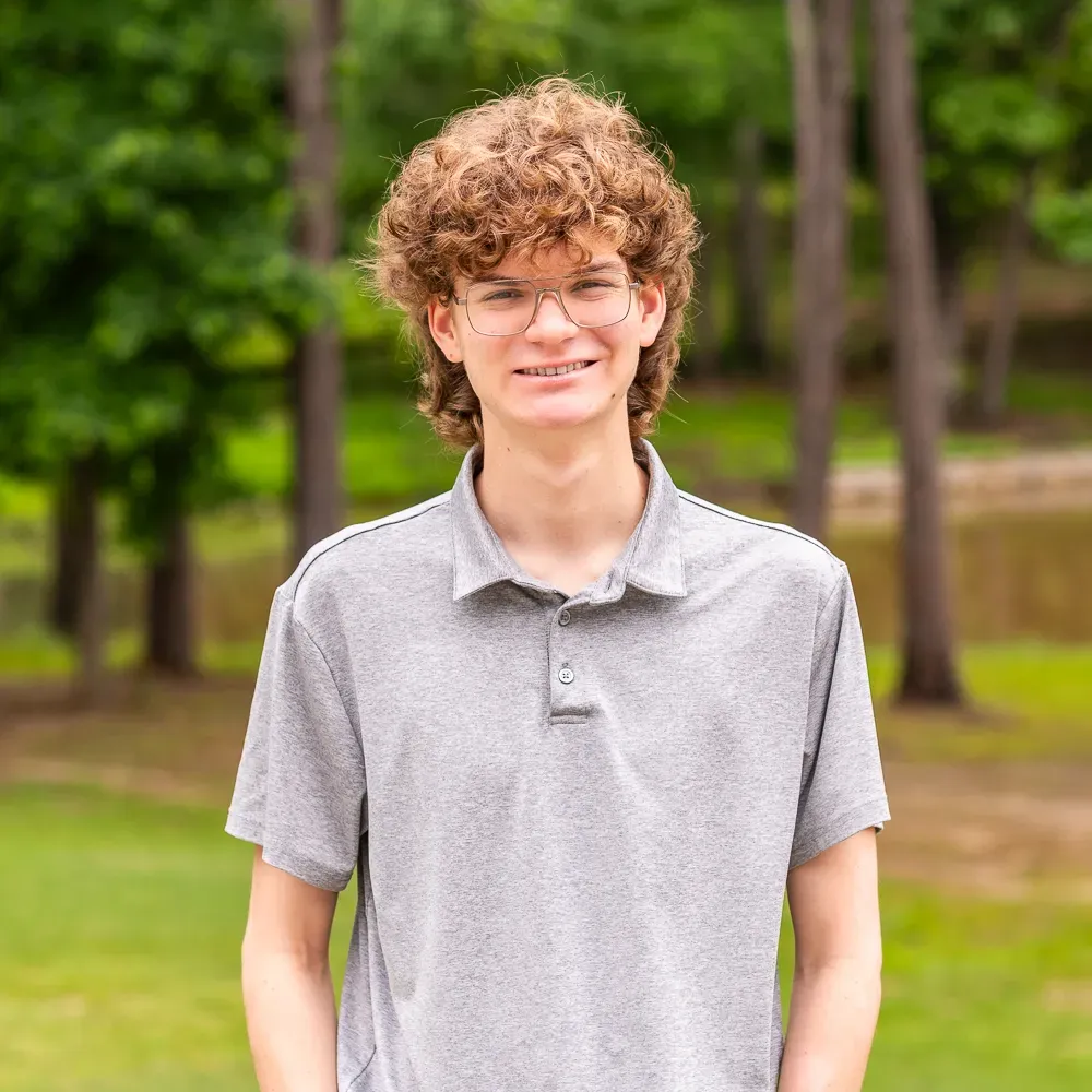 Person with curly hair and glasses wearing a gray polo shirt, standing outdoors in a park.