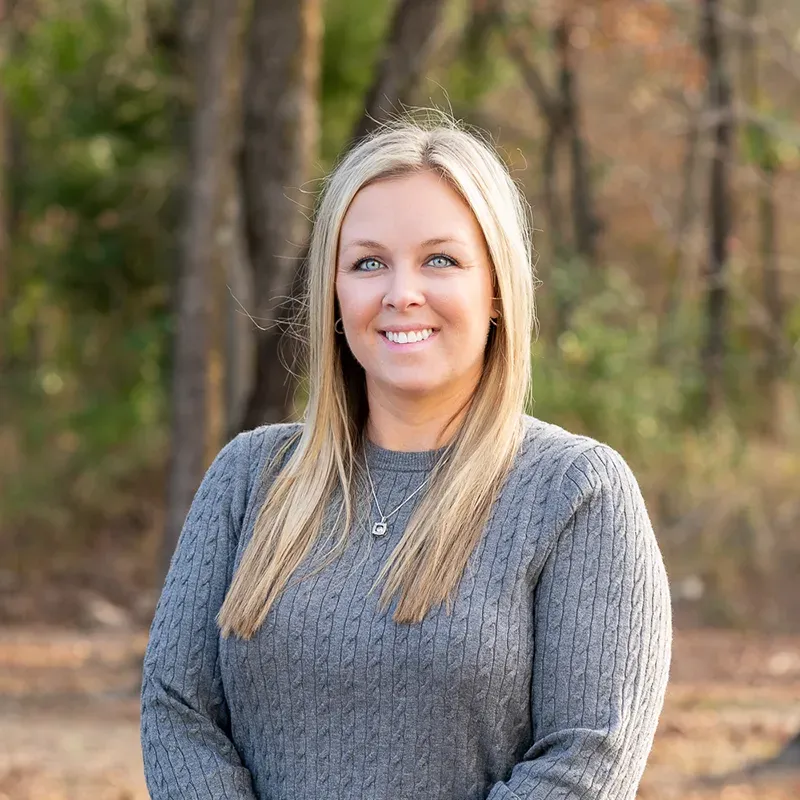 A smiling person with long blonde hair wearing a grey cable-knit sweater, standing outdoors in front of a blurred forest.