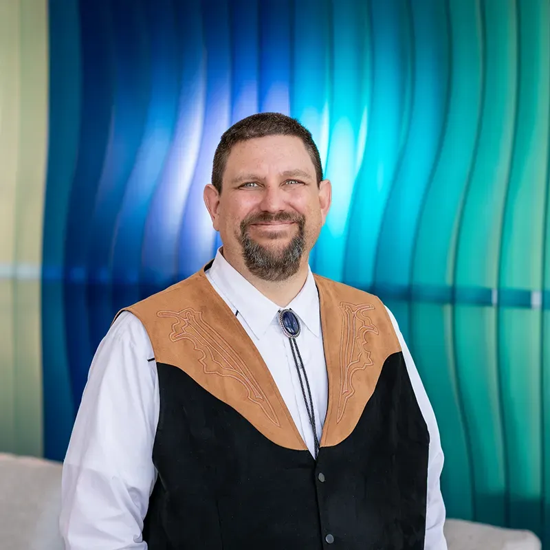 Man in white shirt and bolo tie standing before a blue-green abstract backdrop
