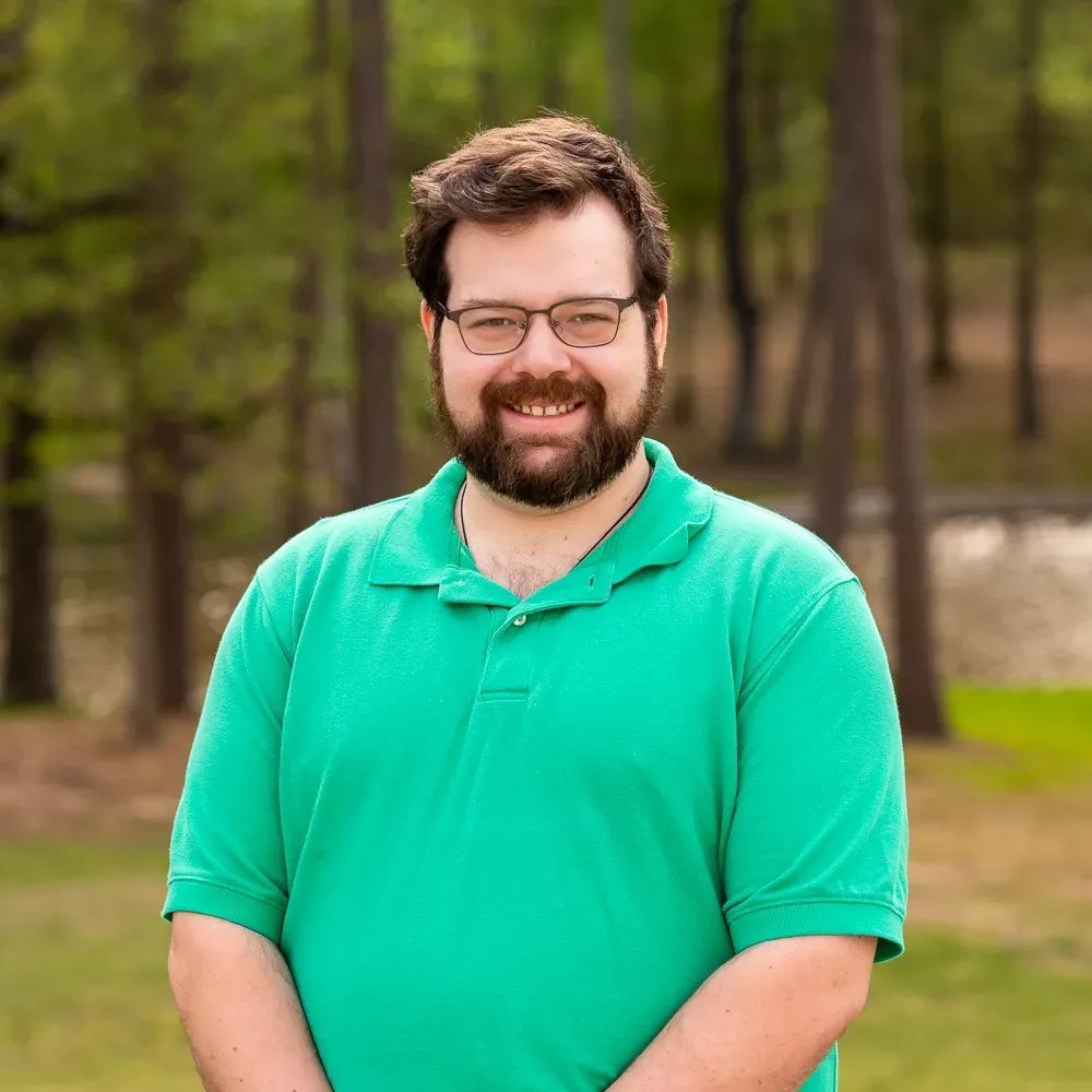 Smiling man in a green polo shirt standing outdoors in a wooded park setting