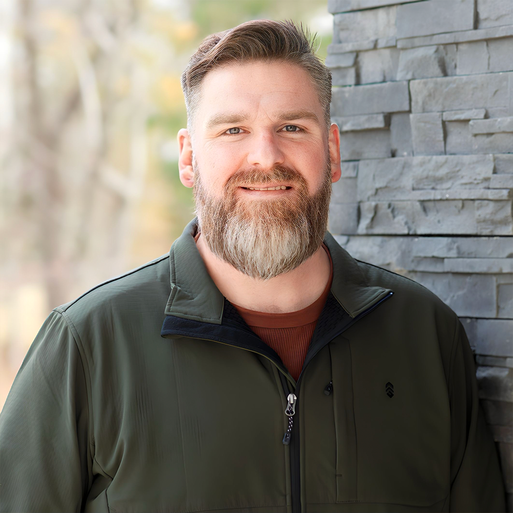 A person with a beard, wearing an olive-green zip-up jacket over a rust-colored shirt, standing against a stone wall.