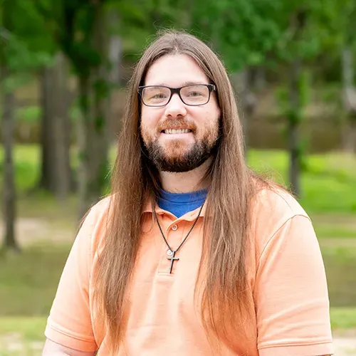 A person with long brown hair and glasses smiles while wearing a peach polo shirt and a cross necklace outdoors.