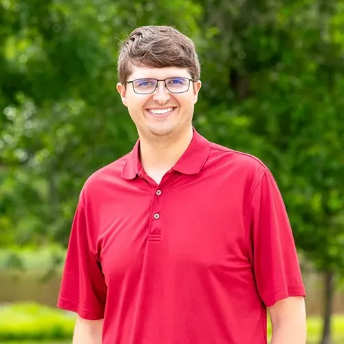 A smiling person wearing glasses and a red polo shirt stands outdoors in front of a blurred green leafy background.