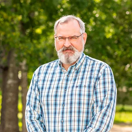 A person with a beard and glasses wearing a blue plaid shirt, standing outdoors against a background of green trees.