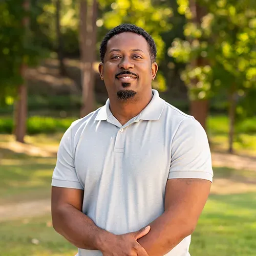 A person with a beard and short hair smiles while wearing a light gray polo shirt in a blurred, sunny park setting.