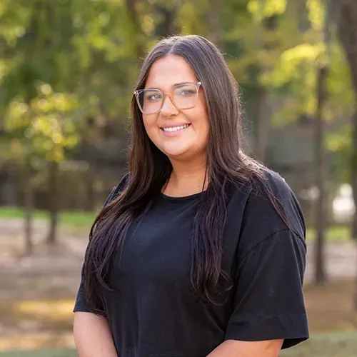 A smiling person with long dark hair and glasses wears a black shirt, posed outdoors against a soft-focus wooded background.
