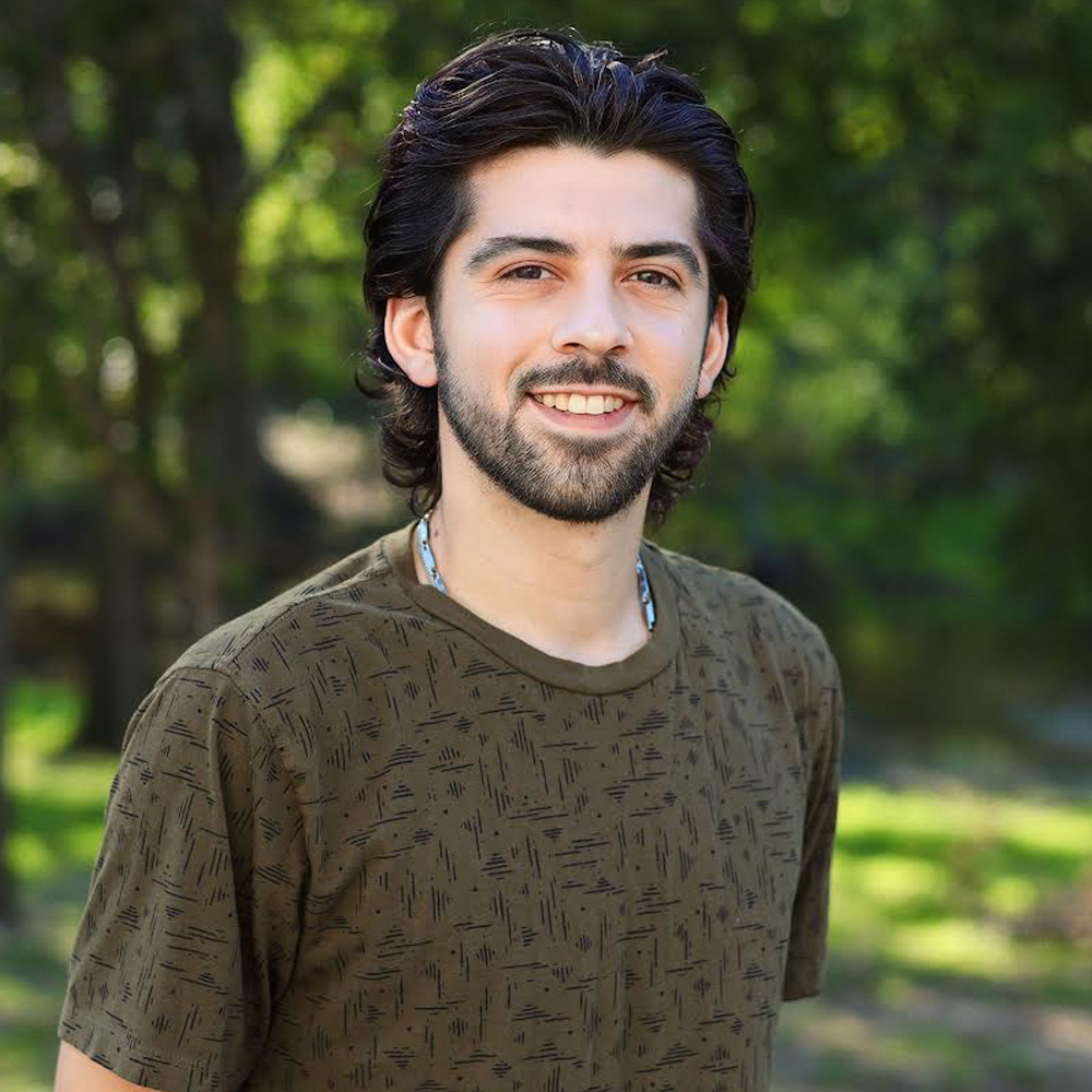 A person with dark, wavy hair and a beard, smiling, wearing a dark patterned t-shirt outdoors with a blurred tree background.