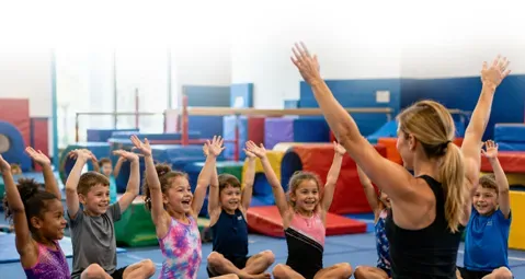 Children in a gymnastics class raising their hands with an instructor in a colorful gym