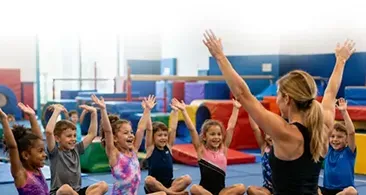 Children in a gymnastics class raising their hands with an instructor in a colorful gym