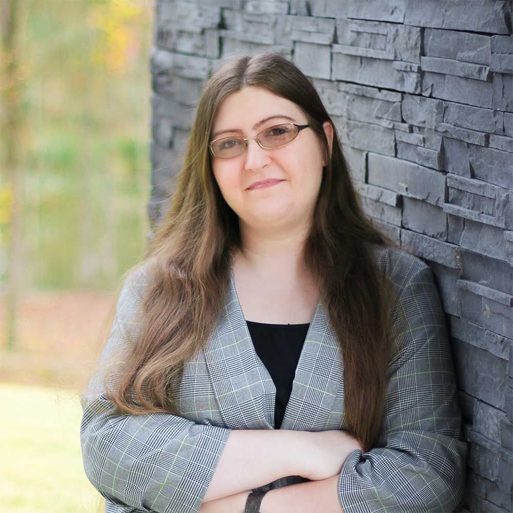 A person with long brown hair and glasses, wearing a grey patterned blazer, stands with arms crossed against a stone wall.