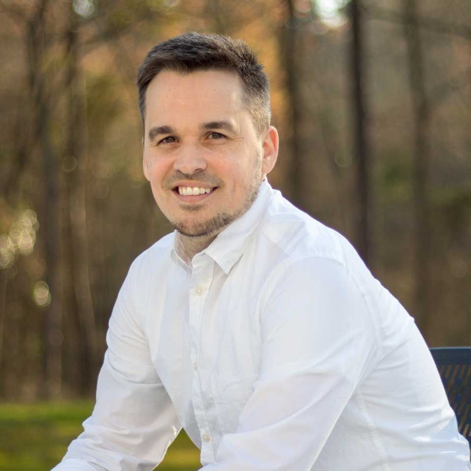 A person with a short beard smiling, wearing a white button-down shirt outdoors with a blurred nature background.