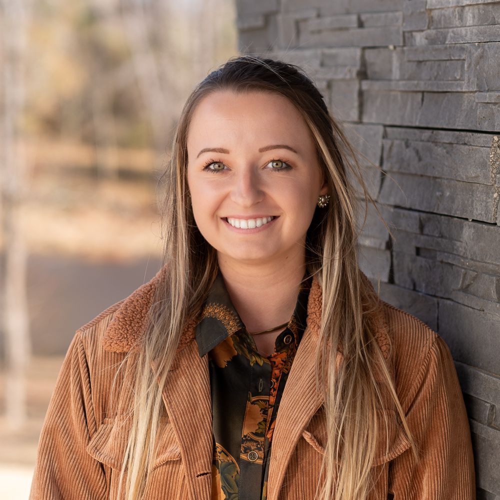 A woman smiling in a tan corduroy jacket against a dark, textured stone wall with a blurred landscape in the background.