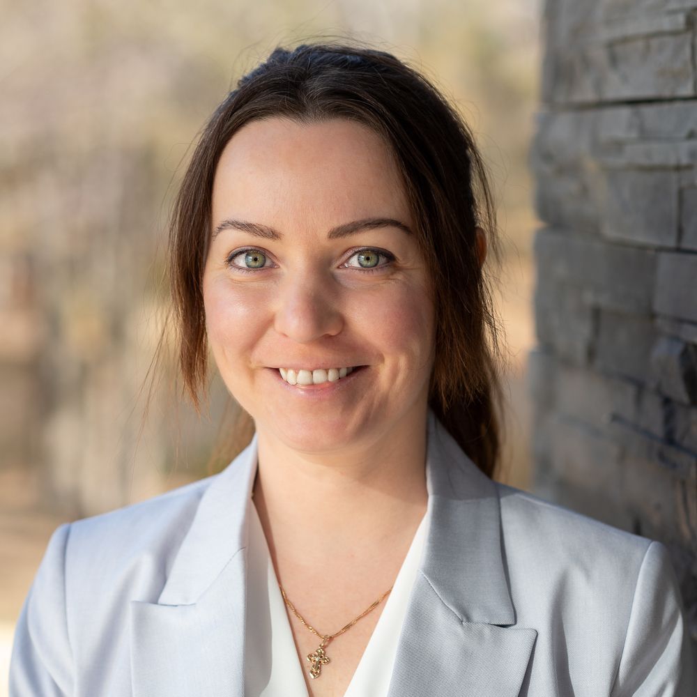 A professional headshot of a smiling person with dark hair, wearing a light gray blazer and a gold necklace.