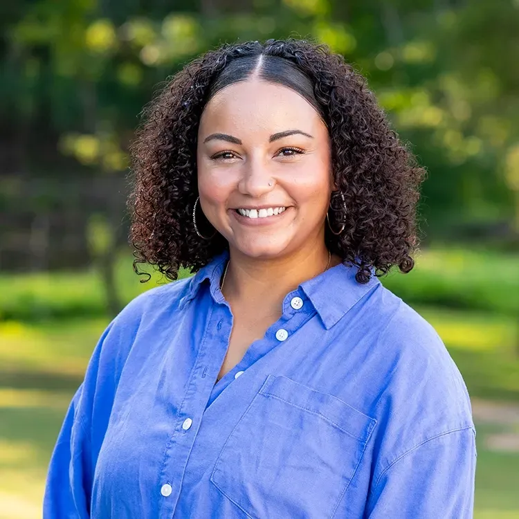 A smiling person with curly dark hair wearing a blue button-down shirt, set against a soft-focus green outdoor background.