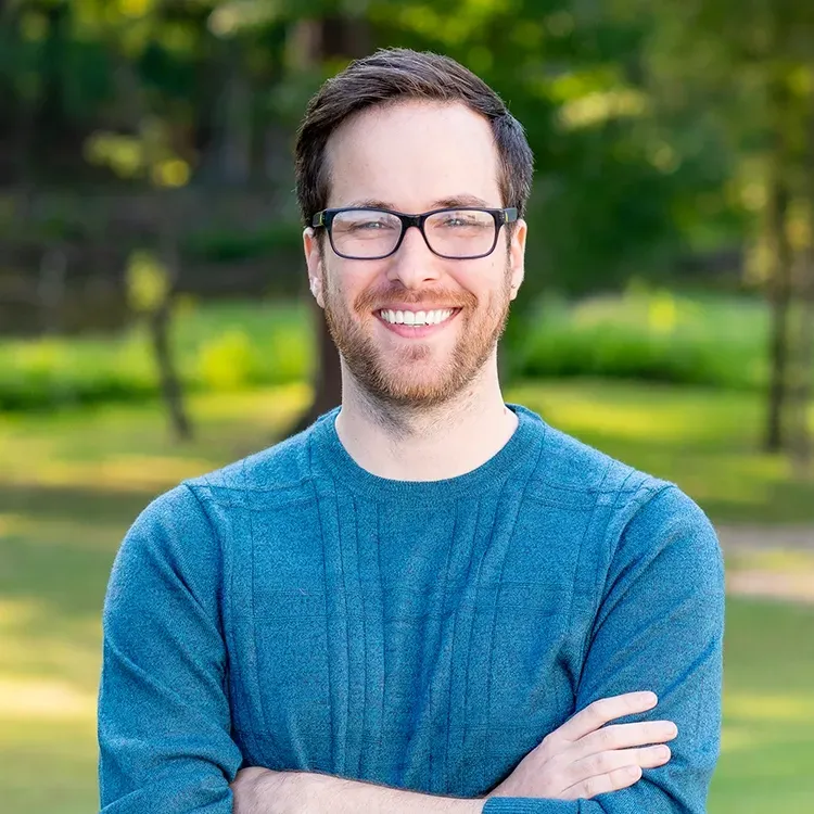 A smiling person with brown hair and glasses, wearing a blue textured sweater, standing with arms crossed outdoors.