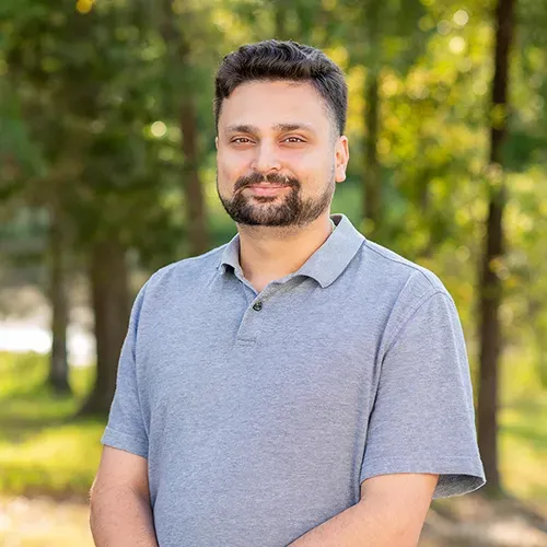 A man with a short beard wearing a grey polo shirt, standing outdoors in front of a soft-focus wooded background.