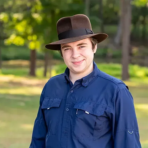 A person wearing a brown fedora and a dark blue long-sleeve button-up shirt, smiling outdoors with a blurred park background.