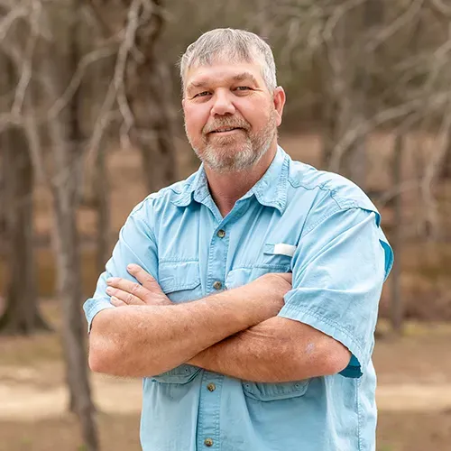 A person in a light blue collared shirt with arms crossed, standing outdoors in front of a blurry, wooded background.