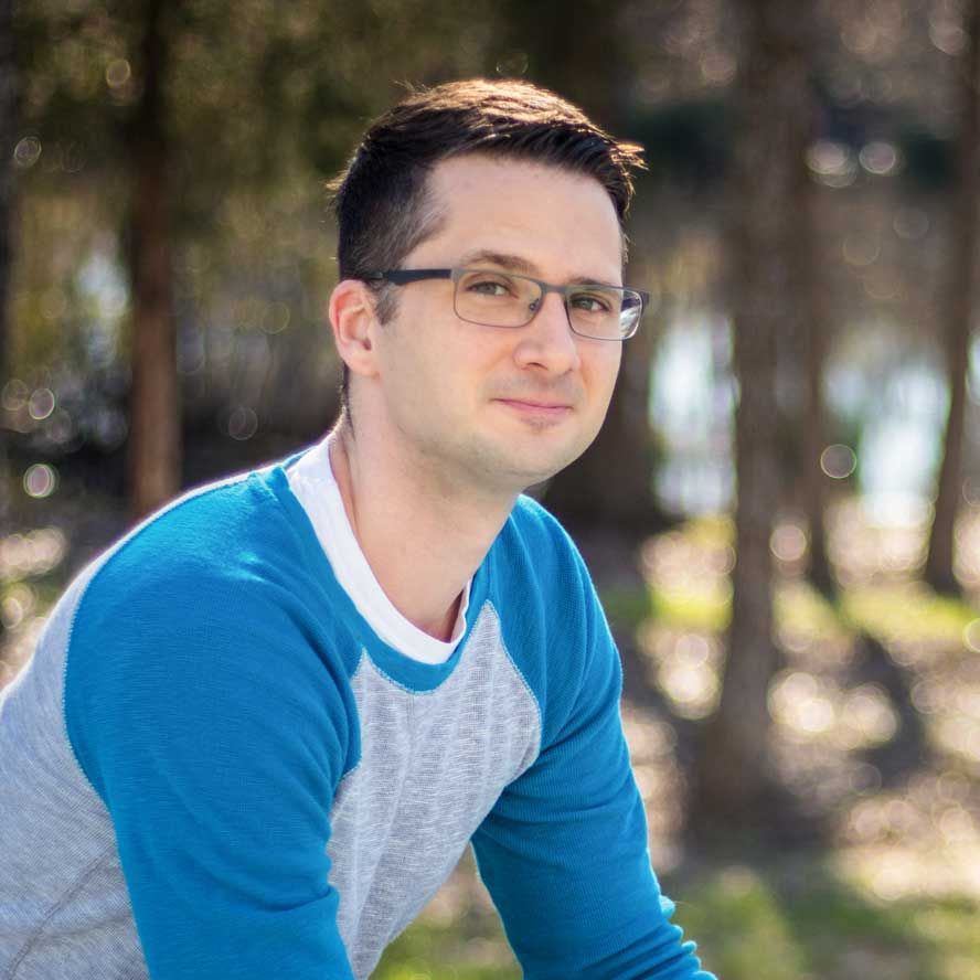 A person wearing glasses and a blue and grey raglan shirt looks at the camera against a blurred outdoor forest background.