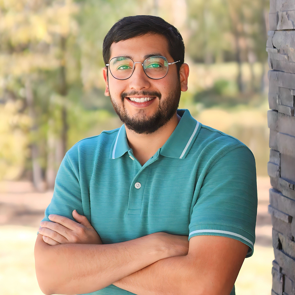 A smiling person with glasses and a beard wears a teal polo shirt, posing with arms crossed against a blurred outdoor background.