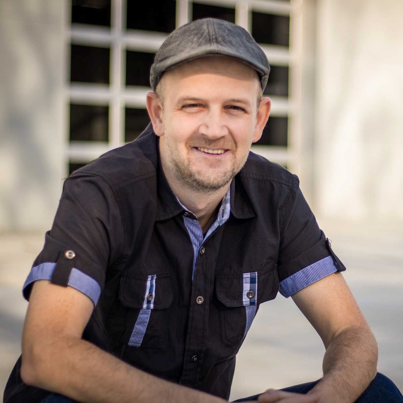 A smiling person wearing a grey flat cap and a dark short-sleeved shirt with blue patterned accents.