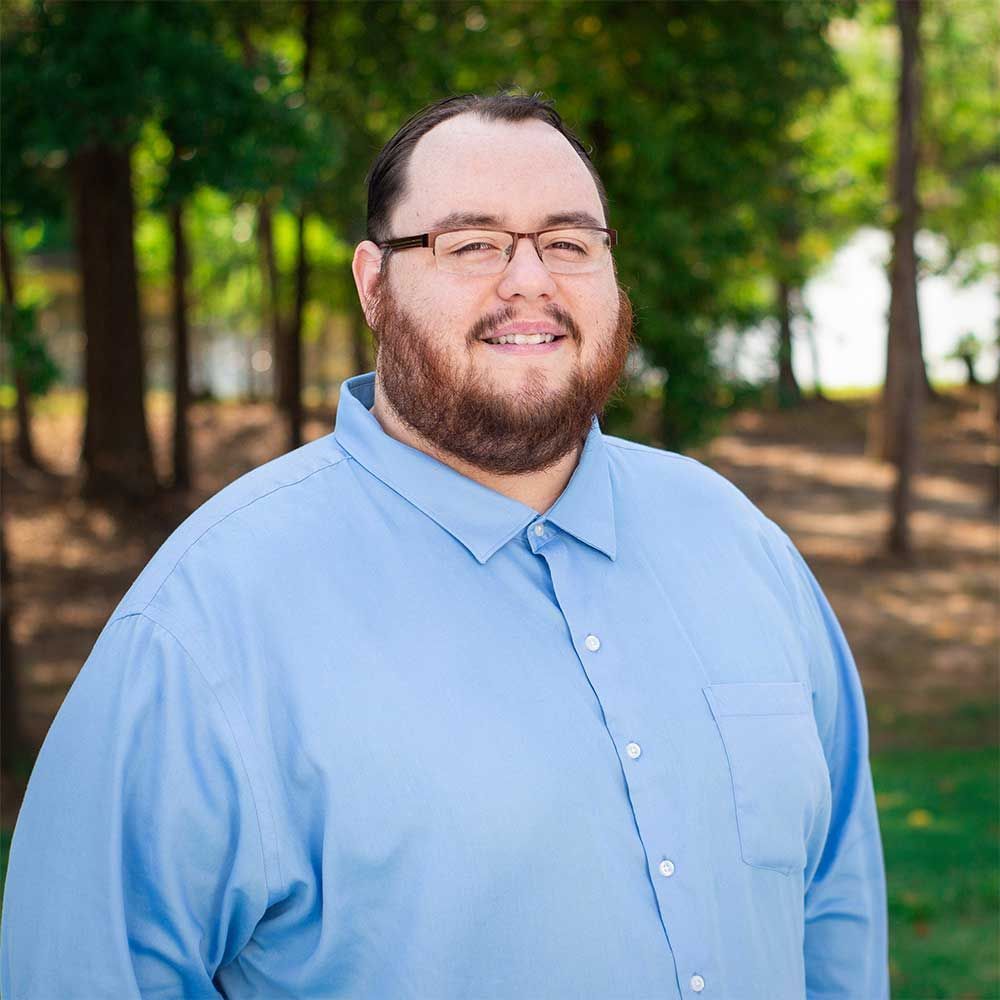 A person with a beard and glasses wearing a light blue button-down shirt, smiling outdoors in front of a wooded area.