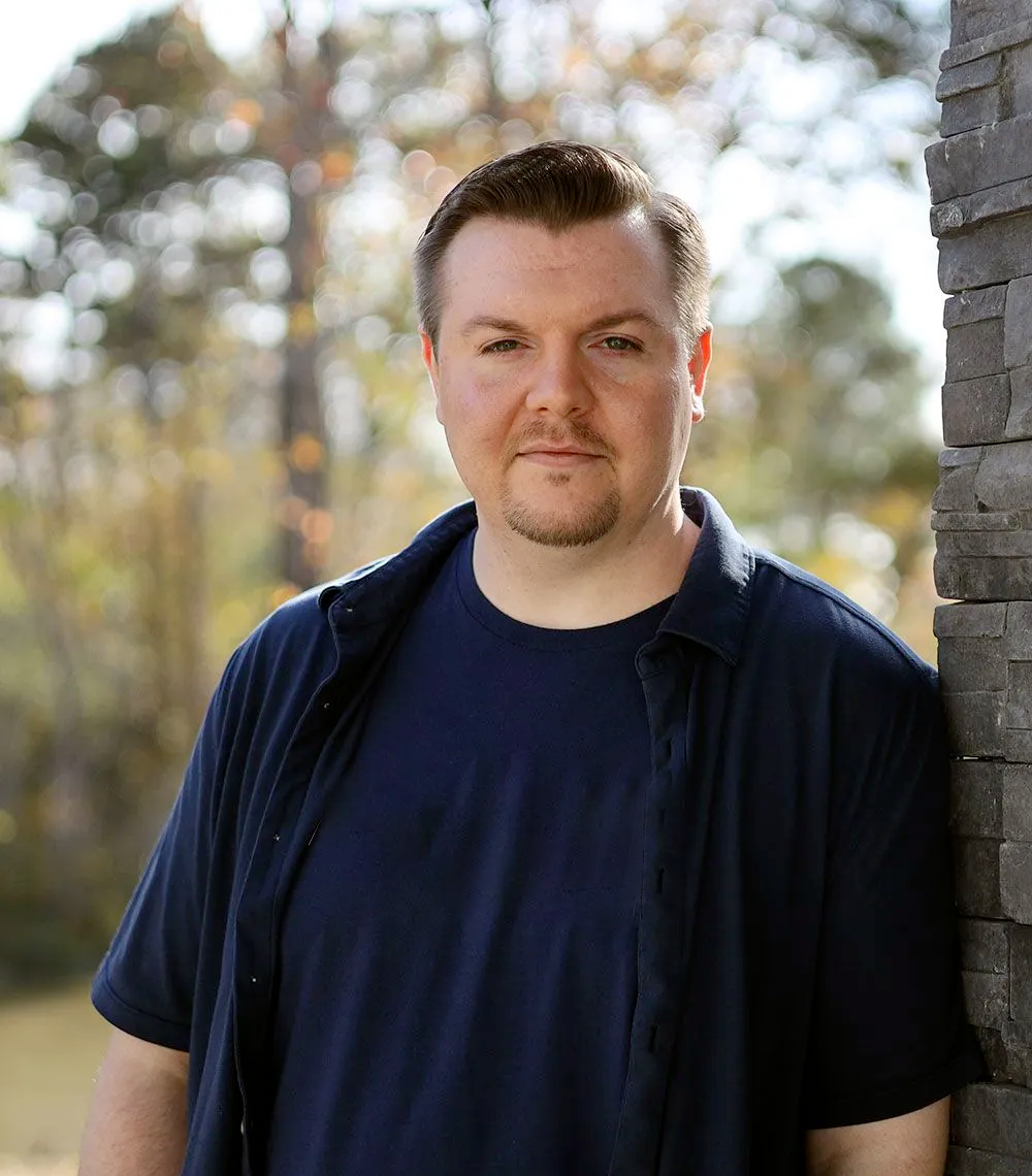 A person with short hair and a goatee stands outdoors, wearing a navy blue shirt and an open dark collared shirt.