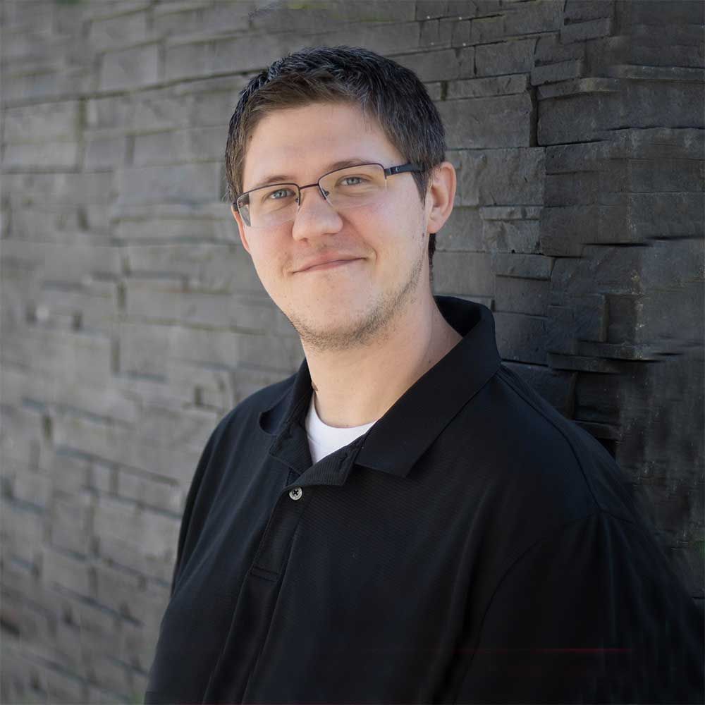 A person with short hair and glasses wearing a black polo shirt, smiling against a dark, textured stone wall.