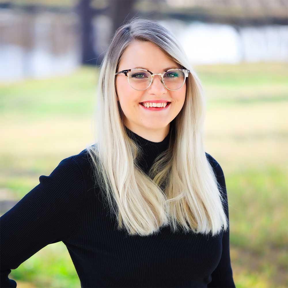 A smiling person with long blonde hair and glasses, wearing a black turtleneck top, standing outdoors in a park setting.
