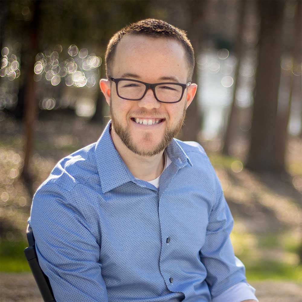 A person with short hair and glasses wearing a blue patterned button-down shirt, smiling outdoors in a wooded area.
