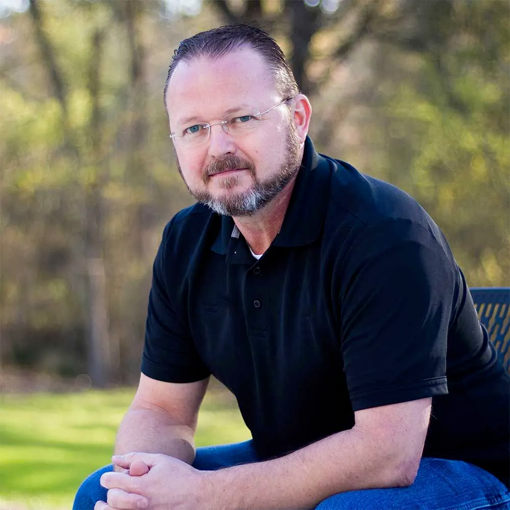 A man with a short beard and glasses wearing a black polo shirt, sitting outdoors with a blurred nature background.