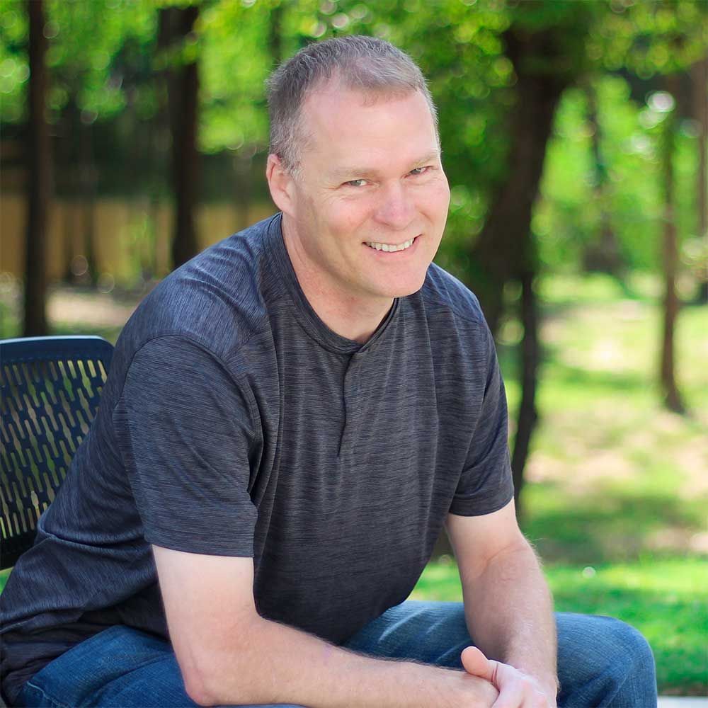A smiling person in a gray t-shirt and blue jeans sits in a chair outdoors with a blurred, green, wooded background.