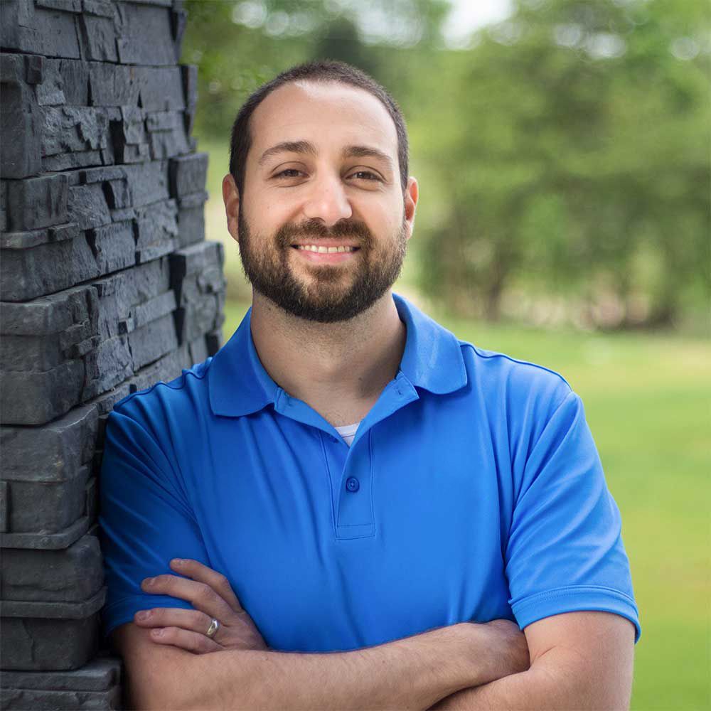 A bearded individual in a blue polo shirt smiles while leaning against a dark stone wall outdoors.
