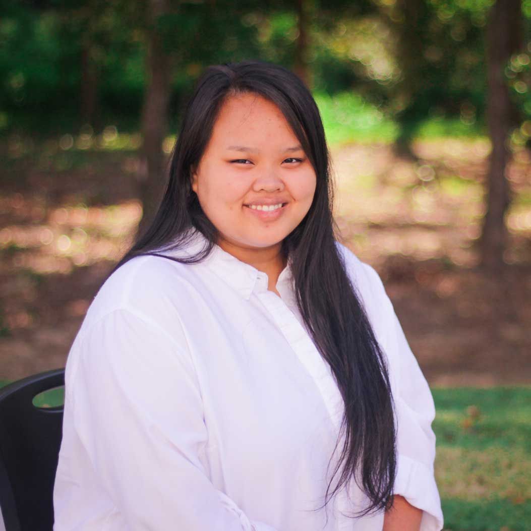 A person with long dark hair and a white button-down shirt smiling in front of a blurred green, outdoor background.