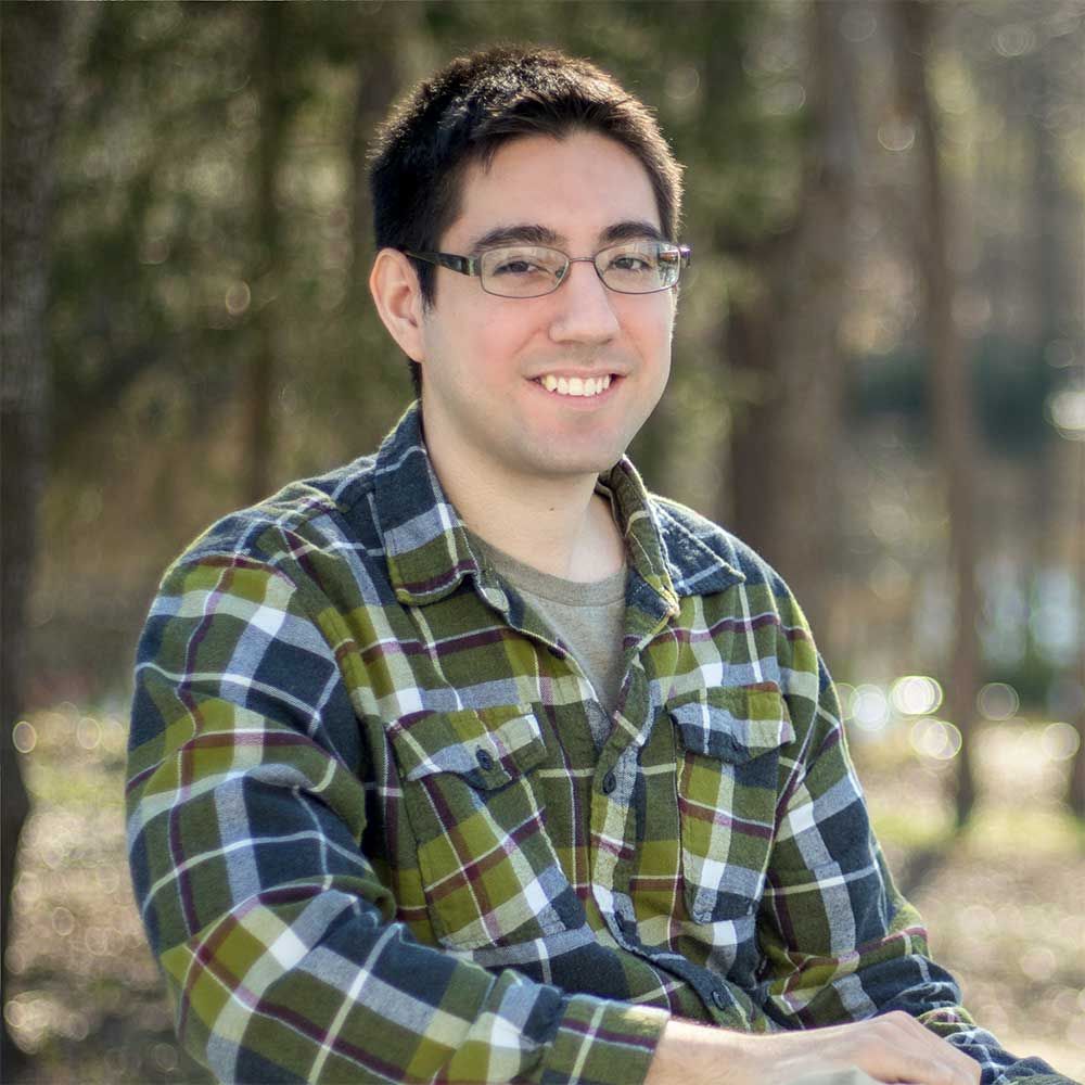 A person with short dark hair and glasses smiles, wearing a green and navy plaid shirt, outdoors in a wooded setting.