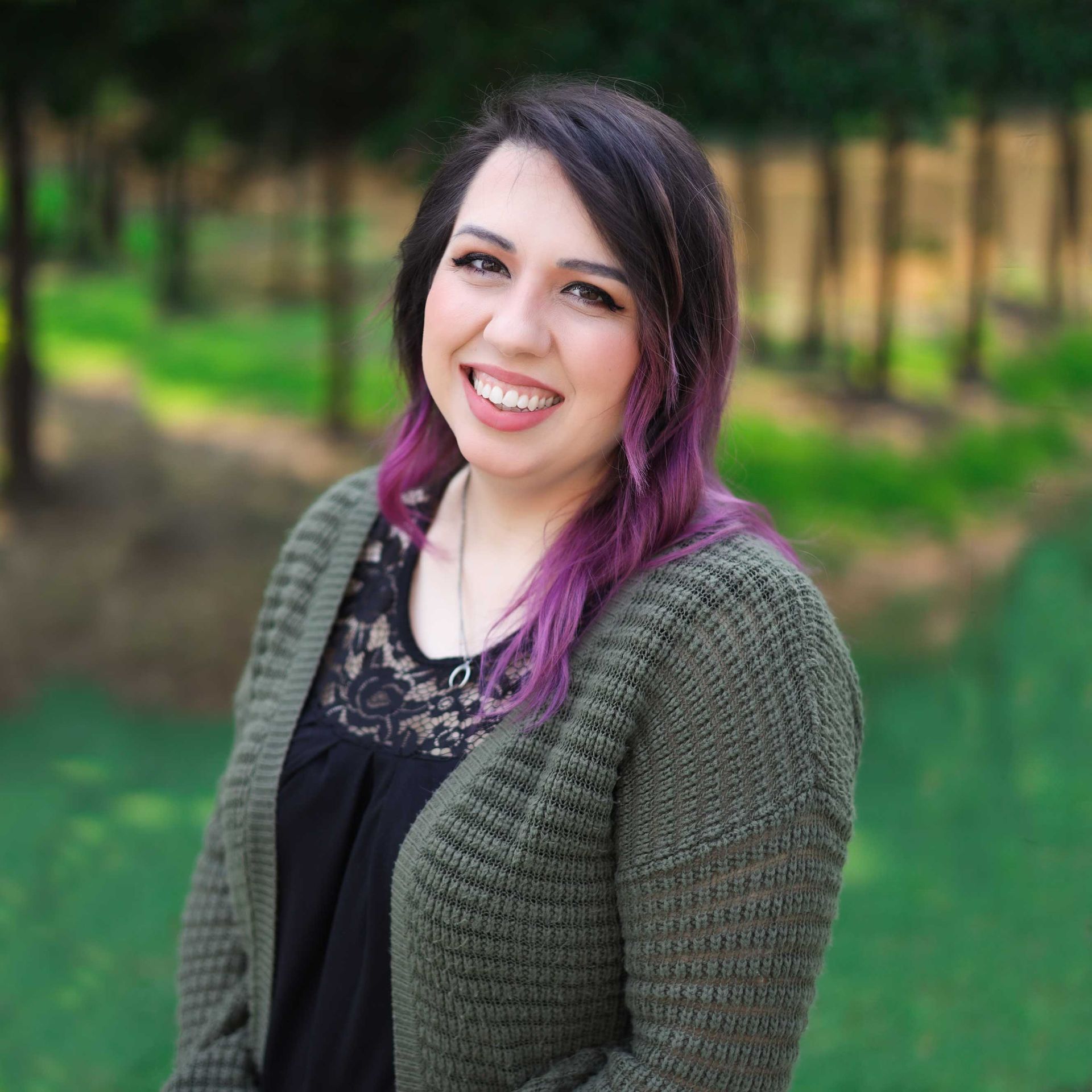 A person with shoulder-length hair with purple highlights smiles in an outdoor setting with trees in the background.