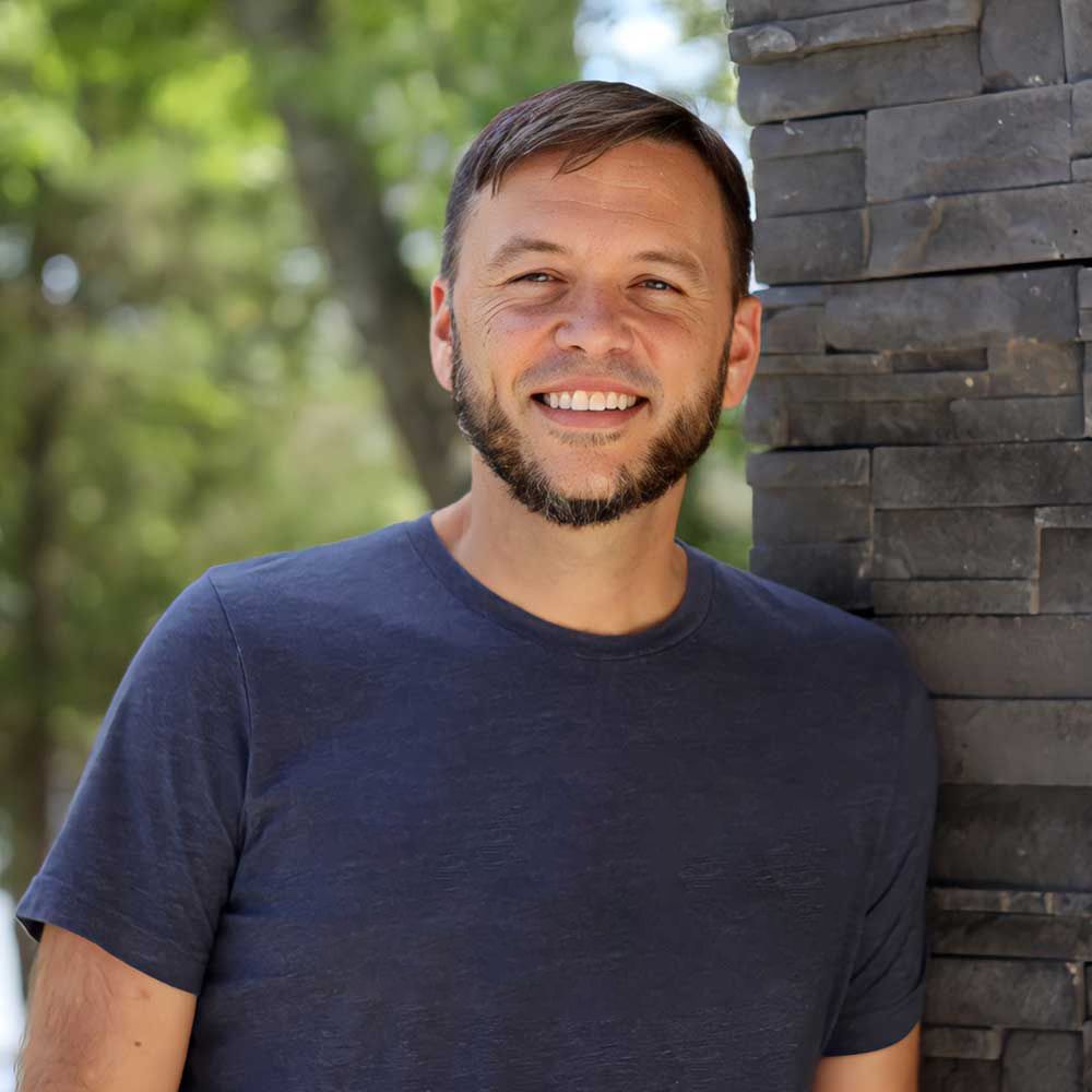 A person with a beard and short hair in a blue t-shirt smiles while leaning against a dark stone wall outdoors.