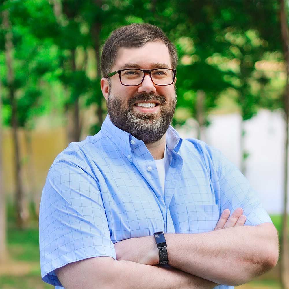 A smiling person with a beard and glasses wears a light blue button-up shirt