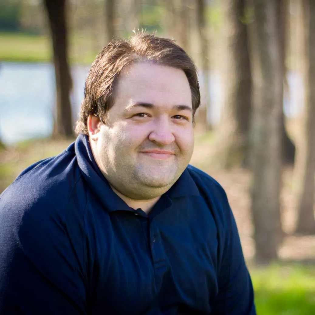 A person in a blue collared shirt smiling softly in front of blurred trees and a water feature.