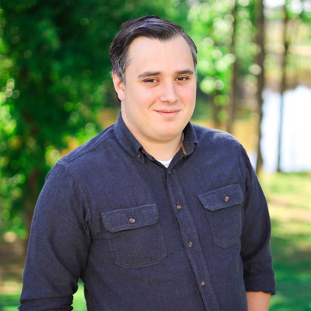 A person with short, dark hair wears a navy blue button-down shirt, standing outdoors with trees in the background.