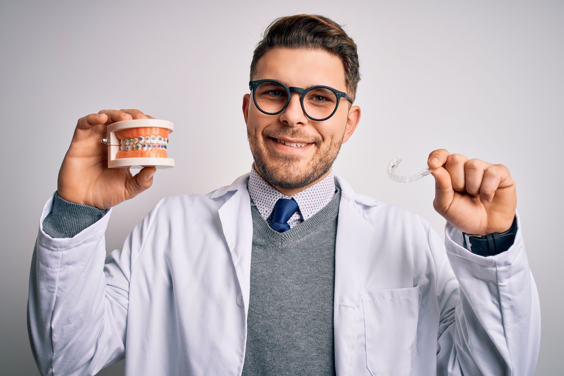 A dentist smiling and holding dentures with braces in one hand and an invisible aligner in the other. A dentist smiling and holding dentures with braces in one hand and an invisible aligner in the other.