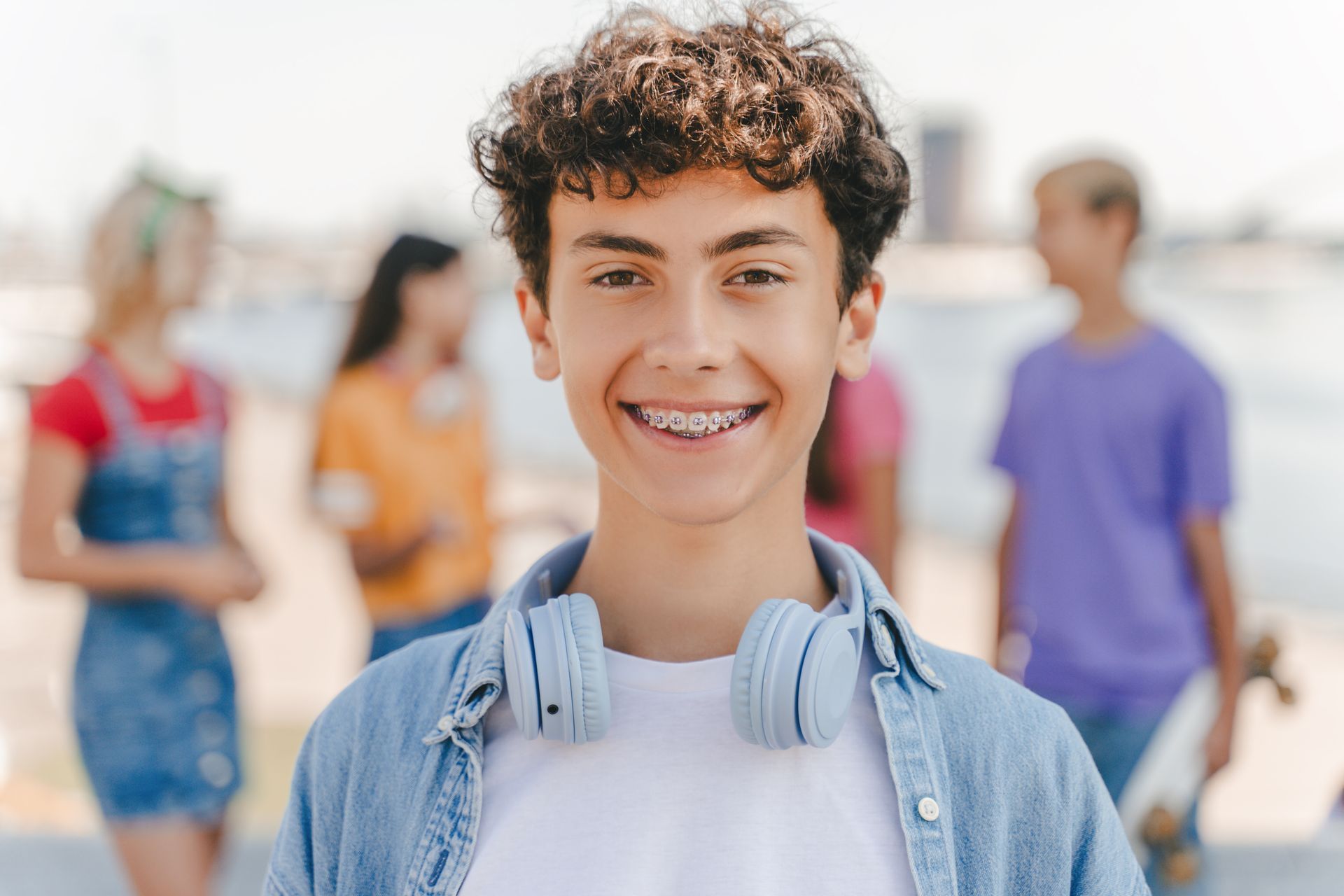 A smiling teenage boy with braces wearing headphones with friends in the background. A smiling teenage boy with braces wearing headphones with friends in the background.