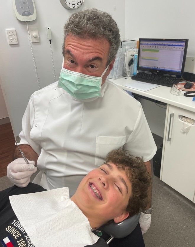 Dentist with a syringe, looking at the camera. Boy in chair smiling. Medical office setting.