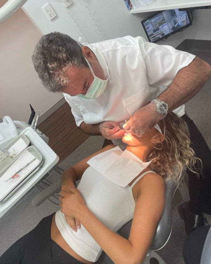 Dentist examining a woman's teeth. He wears a mask and scrubs. The woman is in a chair with a bib.