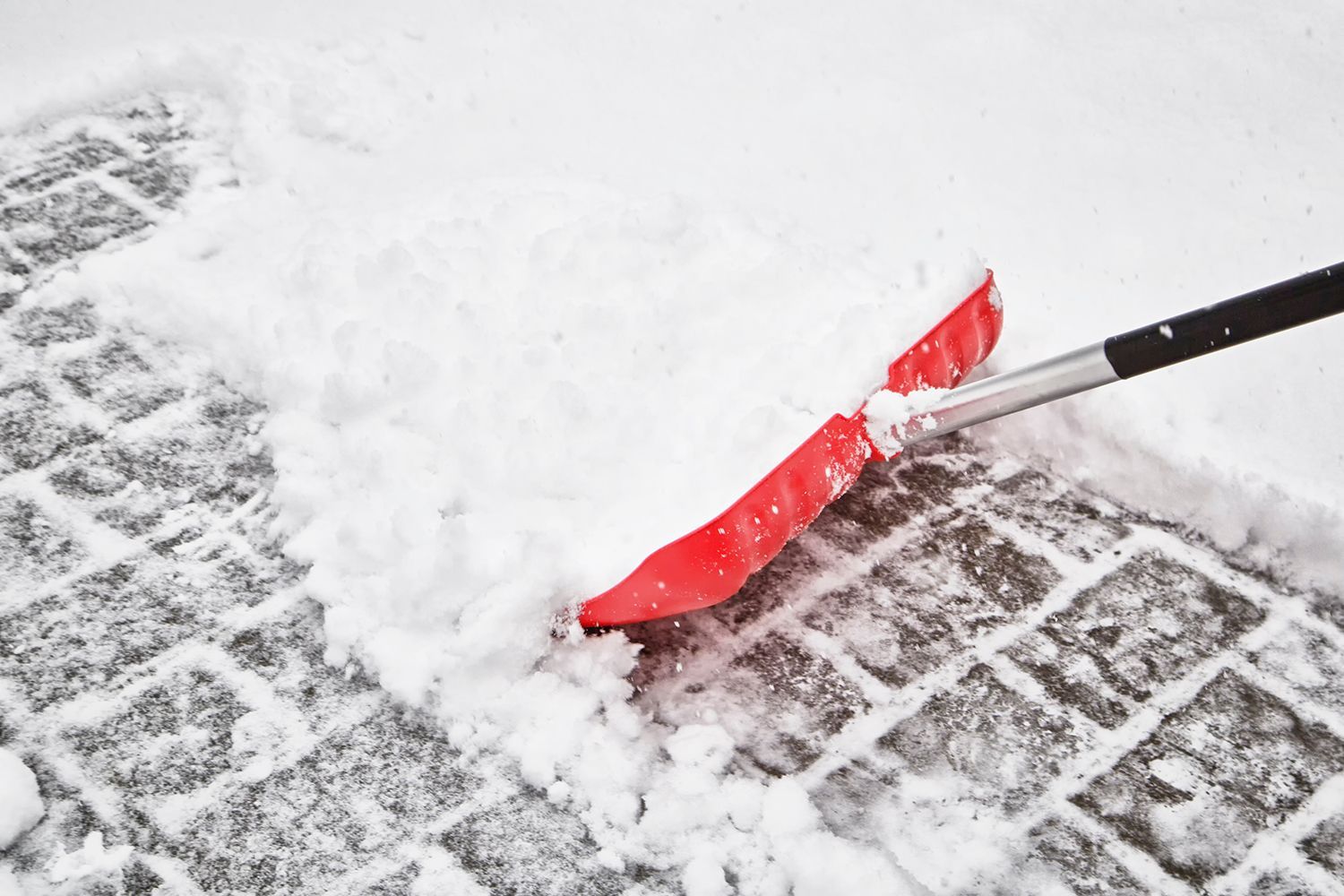 Person shoveling snow with a red snow shovel on a snowy pavement