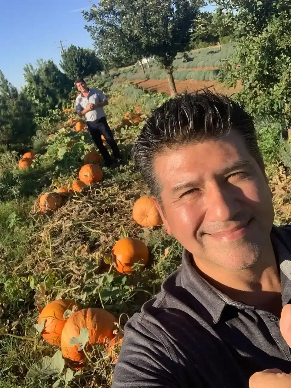 A man taking a selfie in a pumpkin patch with orange pumpkins and another person in the background