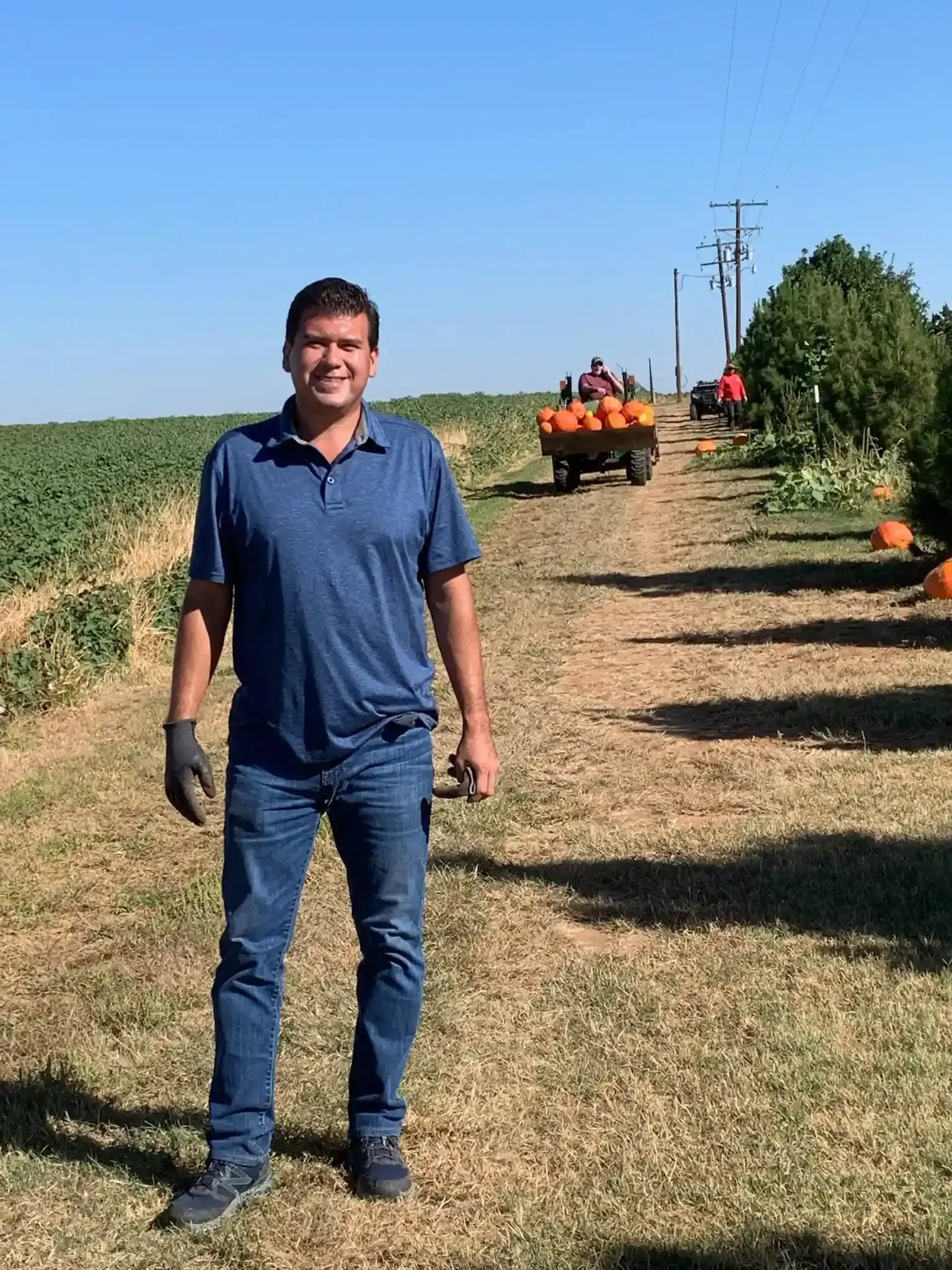 A man standing on a dirt farm path beside crop rows, with a tractor and workers in the background